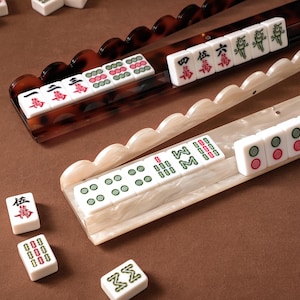 May include: A set of white Mahjong tiles with green and red characters and symbols, arranged on two long racks. One rack is dark brown with a tortoiseshell pattern, and the other is a pearlescent white with a scalloped edge design.