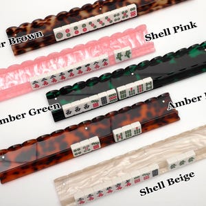 Puede incluir: Cinco soportes para fichas de Mahjong en colores Amber Brown, Shell Pink, Amber Green, Amber Red y Shell Beige. Cada soporte tiene un borde superior festoneado y sostiene una fila de fichas blancas con varios s&iacute;mbolos en rojo, verde y negro.
