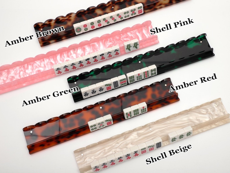 Puede incluir: Cinco soportes para fichas de Mahjong en colores Amber Brown, Shell Pink, Amber Green, Amber Red y Shell Beige. Cada soporte tiene un borde superior festoneado y sostiene una fila de fichas blancas con varios s&iacute;mbolos en rojo, verde y negro.