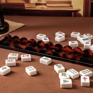 Puede incluir: Un soporte largo para fichas de mahjong con borde festoneado y patr&oacute;n de carey marr&oacute;n oscuro, acompa&ntilde;ado de una colecci&oacute;n de fichas de mahjong rectangulares blancas con varios s&iacute;mbolos y caracteres grabados en verde, rojo y negro dispersos.