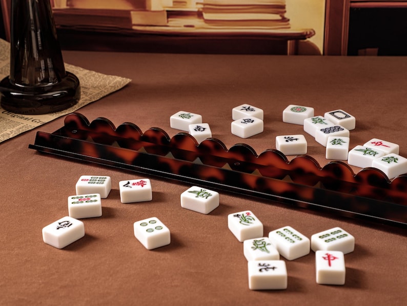 Puede incluir: Un soporte largo para fichas de mahjong con borde festoneado y patr&oacute;n de carey marr&oacute;n oscuro, acompa&ntilde;ado de una colecci&oacute;n de fichas de mahjong rectangulares blancas con varios s&iacute;mbolos y caracteres grabados en verde, rojo y negro dispersos.