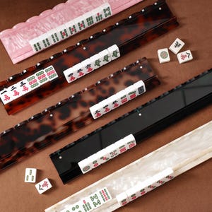 Puede incluir: Cuatro soportes rectangulares largos para fichas de mahjong con patrones en rosa, carey, negro y m&aacute;rmol blanco. Cada soporte presenta un borde festoneado o con muescas para sostener fichas blancas con grabados de caracteres y s&iacute;mbolos.