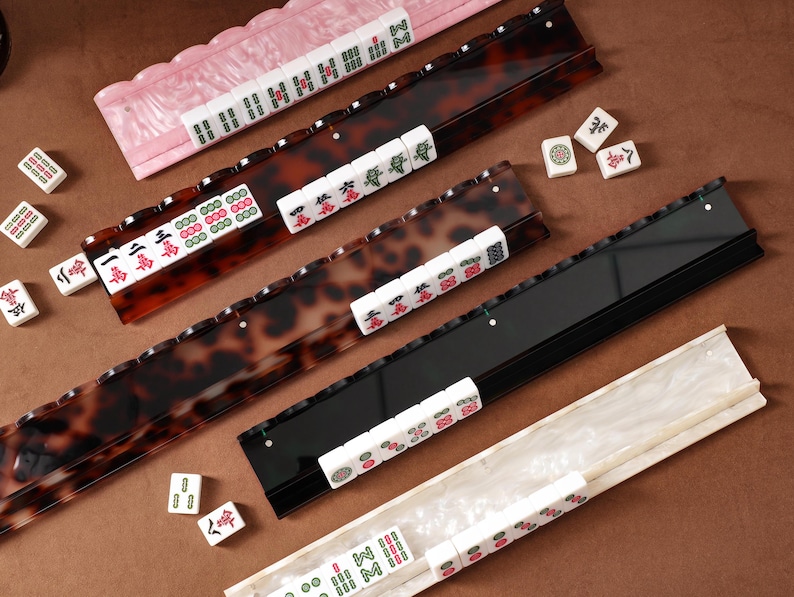Puede incluir: Cuatro soportes rectangulares largos para fichas de mahjong con patrones en rosa, carey, negro y m&aacute;rmol blanco. Cada soporte presenta un borde festoneado o con muescas para sostener fichas blancas con grabados de caracteres y s&iacute;mbolos.