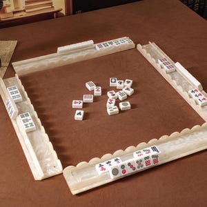 Puede incluir: Un juego de Mahjong que incluye cuatro soportes para fichas blancos con bordes festoneados y acabado marmolado, junto con una colecci&oacute;n de fichas rectangulares blancas marcadas con varios s&iacute;mbolos, caracteres y puntos negros, rojos y verdes.