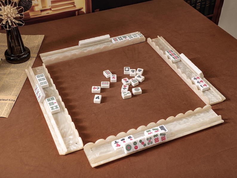 Puede incluir: Un juego de Mahjong que incluye cuatro soportes para fichas blancos con bordes festoneados y acabado marmolado, junto con una colecci&oacute;n de fichas rectangulares blancas marcadas con varios s&iacute;mbolos, caracteres y puntos negros, rojos y verdes.