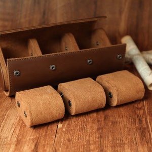 K&ouml;nnte beinhalten: Eine braune Lederuhrbox mit vier F&auml;chern zur Aufbewahrung von Uhren. Die Box ist ge&ouml;ffnet und drei braune Uhrenkissen aus Wildleder sind sichtbar.