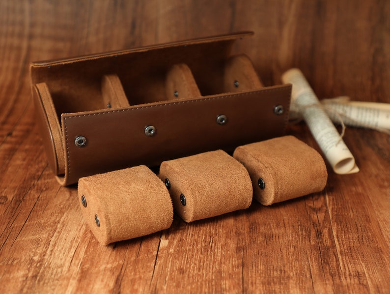 K&ouml;nnte beinhalten: Eine braune Lederuhrbox mit vier F&auml;chern zur Aufbewahrung von Uhren. Die Box ist ge&ouml;ffnet und drei braune Uhrenkissen aus Wildleder sind sichtbar.