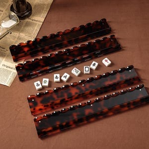 Puede incluir: Cuatro soportes rectangulares para fichas de mahjong con un patr&oacute;n brillante de carey marr&oacute;n y negro, con bordes superiores festoneados. Cinco fichas de mahjong blancas con caracteres chinos negros y rojos est&aacute;n alineadas entre los soportes.