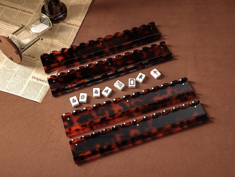 Puede incluir: Cuatro soportes rectangulares para fichas de mahjong con un patr&oacute;n brillante de carey marr&oacute;n y negro, con bordes superiores festoneados. Cinco fichas de mahjong blancas con caracteres chinos negros y rojos est&aacute;n alineadas entre los soportes.