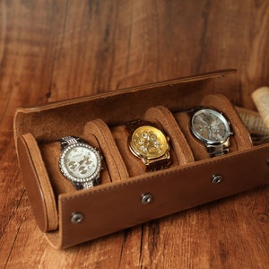 K&ouml;nnte beinhalten: Ein brauner Lederuhrenbeweger mit drei F&auml;chern f&uuml;r Uhren. Der Beweger ist ge&ouml;ffnet und drei Uhren sind sichtbar: eine silberne Uhr mit wei&szlig;em Zifferblatt, eine goldene Uhr mit goldenem Zifferblatt und eine silberne Uhr mit schwarzem Zifferblatt.