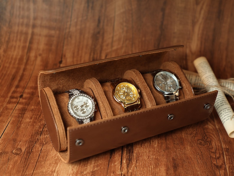 K&ouml;nnte beinhalten: Ein brauner Lederuhrenbeweger mit drei F&auml;chern f&uuml;r Uhren. Der Beweger ist ge&ouml;ffnet und drei Uhren sind sichtbar: eine silberne Uhr mit wei&szlig;em Zifferblatt, eine goldene Uhr mit goldenem Zifferblatt und eine silberne Uhr mit schwarzem Zifferblatt.