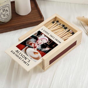 Può includere: Una scatola di fiammiferi in legno con un cassetto scorrevole, contenente fiammiferi e una foto. La foto mostra un bambino a una festa di compleanno, con il testo "I STOLE THIS FROM ALISON'S BIRTHDAY PARTY".