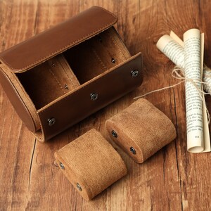 K&ouml;nnte beinhalten: Eine zylindrische Uhrenbox aus braunem Leder mit zwei F&auml;chern, die mit braunem Wildleder ausgekleidet sind. Die Box ist ge&ouml;ffnet und die F&auml;cher sind leer. Zwei zus&auml;tzliche Uhrenkissen aus braunem Wildleder sind ebenfalls zu sehen.