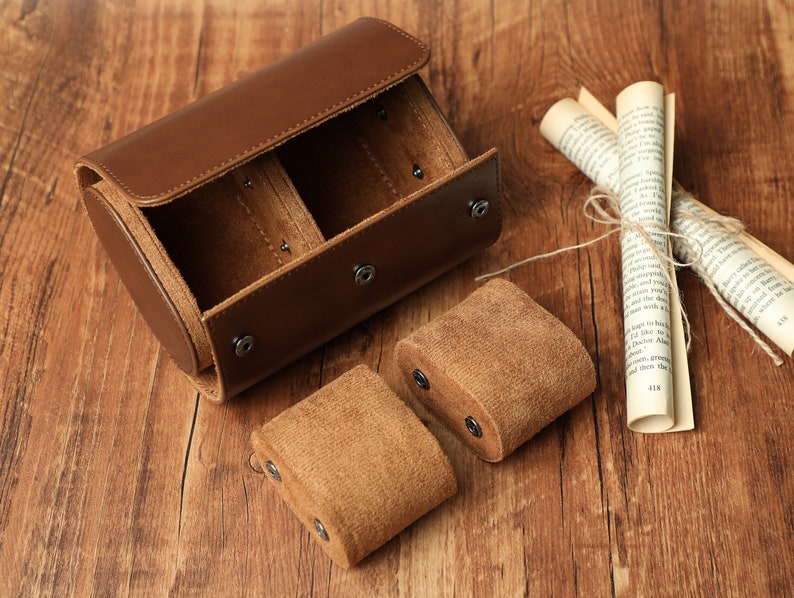 K&ouml;nnte beinhalten: Eine zylindrische Uhrenbox aus braunem Leder mit zwei F&auml;chern, die mit braunem Wildleder ausgekleidet sind. Die Box ist ge&ouml;ffnet und die F&auml;cher sind leer. Zwei zus&auml;tzliche Uhrenkissen aus braunem Wildleder sind ebenfalls zu sehen.