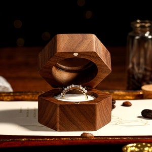 Op de afbeelding: Een zeshoekige houten ringendoos, open om een ​​diamanten ring te laten zien. De doos is gemaakt van donker hout en heeft een gladde afwerking.