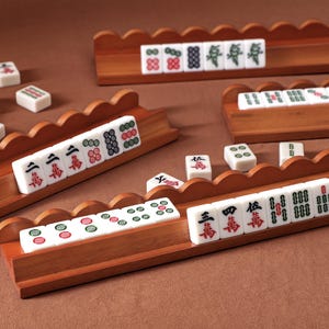 Könnte beinhalten: Ein Set aus vier hölzernen Mahjong-Fliesenhaltern mit gewelltem Rand, die jeweils eine Reihe weißer, rechteckiger Spielsteine mit verschiedenen eingravierten roten, grünen und schwarzen Symbolen, Schriftzeichen und Zahlenmustern halten.