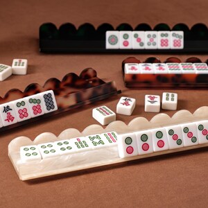 Puede incluir: Una colecci&oacute;n de fichas de Mahjong dispuestas en tres soportes festoneados distintos. Un soporte es negro, otro es de carey marr&oacute;n y otro es de m&aacute;rmol blanco. Las fichas rectangulares blancas presentan varios s&iacute;mbolos y n&uacute;meros en rojo, verde y negro.