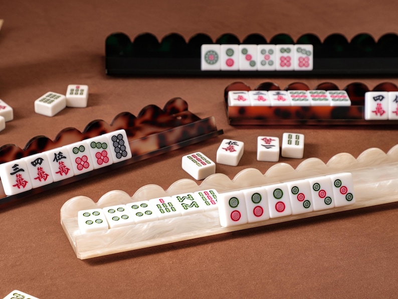 Puede incluir: Una colecci&oacute;n de fichas de Mahjong dispuestas en tres soportes festoneados distintos. Un soporte es negro, otro es de carey marr&oacute;n y otro es de m&aacute;rmol blanco. Las fichas rectangulares blancas presentan varios s&iacute;mbolos y n&uacute;meros en rojo, verde y negro.