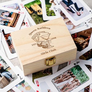 May include: A light wooden box with a gold clasp, surrounded by playing cards featuring photographs. The box is engraved with the words "Queen, Kathleen", "Girls Club", and "1998", along with playing card, martini glass, and dice illustrations.