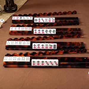 Puede incluir: Cuatro soportes rectangulares y largos para fichas de Mahjong con un patr&oacute;n de carey marr&oacute;n oscuro y bordes superiores festoneados. Cada soporte sostiene una fila de fichas blancas con varios s&iacute;mbolos, caracteres y dise&ntilde;os num&eacute;ricos grabados.