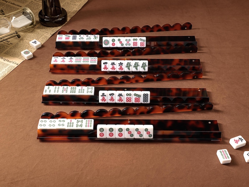 Puede incluir: Cuatro soportes rectangulares y largos para fichas de Mahjong con un patr&oacute;n de carey marr&oacute;n oscuro y bordes superiores festoneados. Cada soporte sostiene una fila de fichas blancas con varios s&iacute;mbolos, caracteres y dise&ntilde;os num&eacute;ricos grabados.