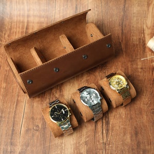 K&ouml;nnte beinhalten: Eine braune Leder-Uhrenrolle mit drei F&auml;chern zum Aufbewahren von Uhren. Drei Uhren werden auf der Rolle pr&auml;sentiert, eine mit schwarzem Zifferblatt, eine mit goldenem Zifferblatt und eine mit silbernem Zifferblatt.