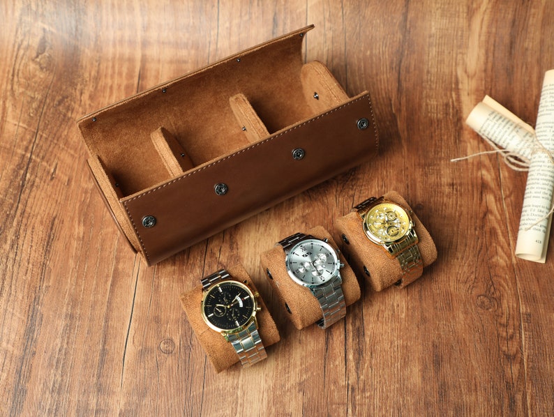 K&ouml;nnte beinhalten: Eine braune Leder-Uhrenrolle mit drei F&auml;chern zum Aufbewahren von Uhren. Drei Uhren werden auf der Rolle pr&auml;sentiert, eine mit schwarzem Zifferblatt, eine mit goldenem Zifferblatt und eine mit silbernem Zifferblatt.