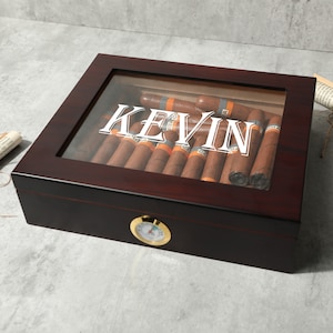 Könnte beinhalten: Eine dunkelbraune Holzkistchen für Zigarren mit Glasdeckel. Die Kiste hat ein Hygrometer an der Vorderseite und den Namen "KEVIN" auf dem Deckel graviert. Die Kiste ist mit Zigarren gefüllt.