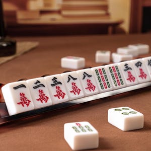 Puede incluir: Un juego de fichas de Mahjong blancas con caracteres chinos en negro y rojo y s&iacute;mbolos verdes, dispuestas en fila sobre un soporte marr&oacute;n con patr&oacute;n de carey, con fichas sueltas esparcidas sobre una superficie marr&oacute;n.