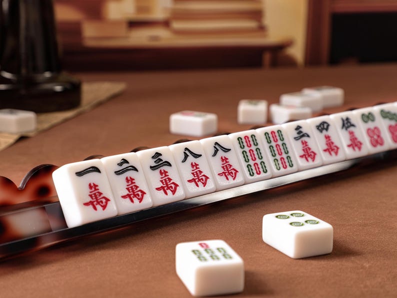 Puede incluir: Un juego de fichas de Mahjong blancas con caracteres chinos en negro y rojo y s&iacute;mbolos verdes, dispuestas en fila sobre un soporte marr&oacute;n con patr&oacute;n de carey, con fichas sueltas esparcidas sobre una superficie marr&oacute;n.