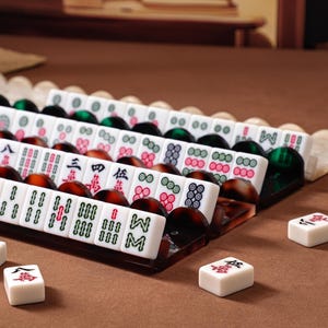 Puede incluir: Un juego de fichas de Mahjong blancas con s&iacute;mbolos grabados en rojo, verde y negro, dispuestas en filas sobre soportes de acr&iacute;lico marr&oacute;n oscuro y verde transl&uacute;cido. Varias fichas individuales est&aacute;n colocadas por separado frente a los soportes.
