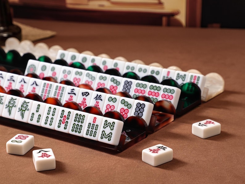 Puede incluir: Un juego de fichas de Mahjong blancas con s&iacute;mbolos grabados en rojo, verde y negro, dispuestas en filas sobre soportes de acr&iacute;lico marr&oacute;n oscuro y verde transl&uacute;cido. Varias fichas individuales est&aacute;n colocadas por separado frente a los soportes.