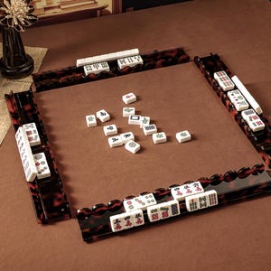 Puede incluir: Un juego de Mahjong con fichas blancas que presentan caracteres y s&iacute;mbolos en rojo, verde y negro. El conjunto incluye cuatro soportes para fichas con patr&oacute;n de carey marr&oacute;n oscuro dispuestos en cuadrado, con fichas dispersas en el centro.