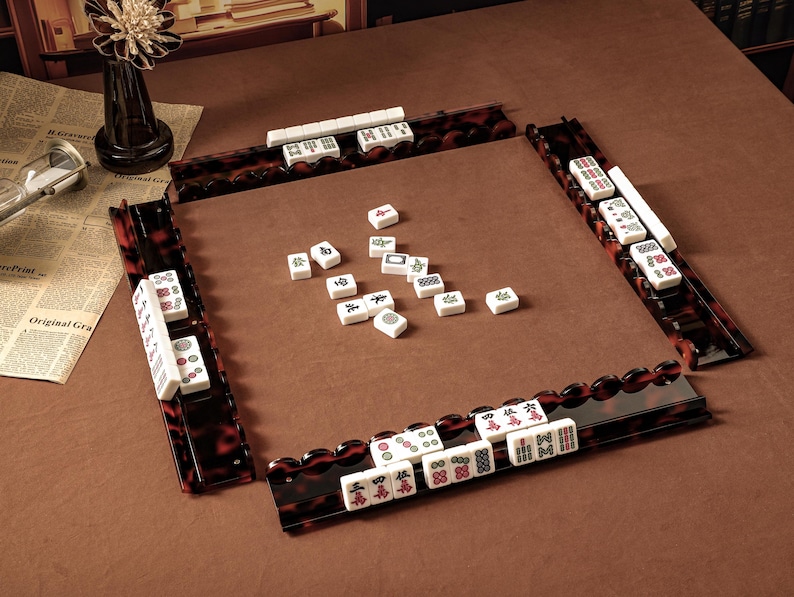 Puede incluir: Un juego de Mahjong con fichas blancas que presentan caracteres y s&iacute;mbolos en rojo, verde y negro. El conjunto incluye cuatro soportes para fichas con patr&oacute;n de carey marr&oacute;n oscuro dispuestos en cuadrado, con fichas dispersas en el centro.