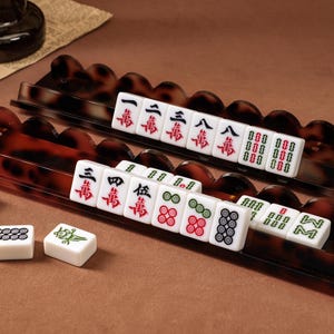 Könnte beinhalten: Zwei lange Mahjong-Fliesenhalter mit gewelltem Rand und braunem Schildpattmuster. Die Halter enthalten weiße Mahjong-Steine mit roten, grünen und schwarzen chinesischen Schriftzeichen und Symbolen. Drei weitere lose Spielsteine liegen daneben.