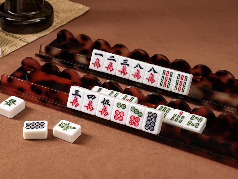 Puede incluir: Dos soportes largos para fichas de mahjong con bordes festoneados y patr&oacute;n de carey marr&oacute;n. Los soportes sostienen fichas blancas con caracteres y s&iacute;mbolos chinos en rojo, verde y negro. Tres fichas sueltas adicionales se encuentran cerca.