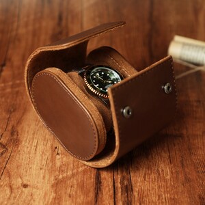 K&ouml;nnte beinhalten: Eine braune Lederuhrenrolle mit einer Uhr darin. Die Uhr hat ein schwarzes Zifferblatt mit wei&szlig;en Markierungen und einer goldenen L&uuml;nette.