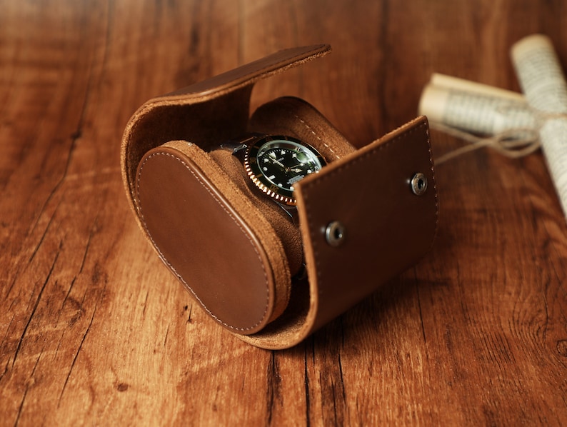 K&ouml;nnte beinhalten: Eine braune Lederuhrenrolle mit einer Uhr darin. Die Uhr hat ein schwarzes Zifferblatt mit wei&szlig;en Markierungen und einer goldenen L&uuml;nette.