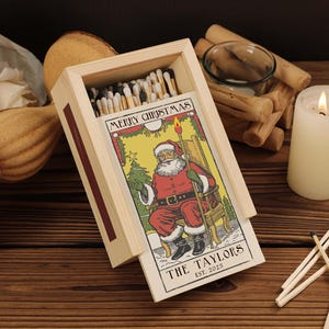 Puede incluir: Caja de cerillas de madera con un diseño navideño. La caja presenta una colorida ilustración de Papá Noel y el texto "MERRY CHRISTMAS". La parte inferior de la caja dice "THE TAYLORS EST. 2025". Una vela encendida y cerillas están al lado de la caja.