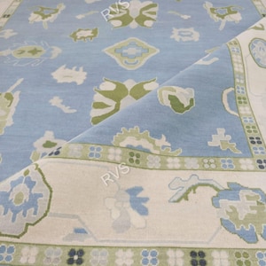 May include: A light blue area rug featuring a repeating pattern of stylized green and white floral motifs. The rug has a wide cream-colored border with a geometric green and blue design and a fringe edge. The text RVS is visible on the rug surface.