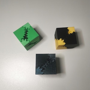 Puede incluir: Tres cubos impresos en 3D con engranajes que se engranan. Los cubos son verde, negro y azul oscuro.