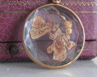 Peut inclure: Un pendentif doré avec un médaillon en verre transparent contenant une fleur séchée pressée. Le médaillon est facetté et de forme arrondie.