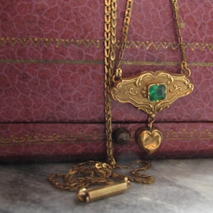 May include: A gold-toned necklace with an ornate pendant featuring a square emerald-green stone and a heart-shaped charm. The necklace is displayed against a textured, reddish-purple background.