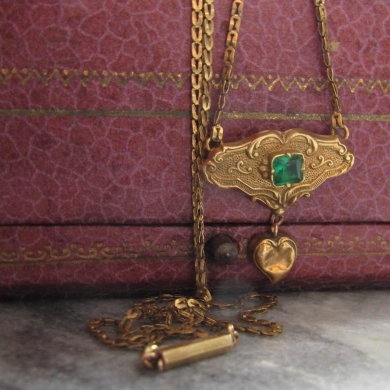 May include: A gold-toned necklace with an ornate pendant featuring a square emerald-green stone and a heart-shaped charm. The necklace is displayed against a textured, reddish-purple background.