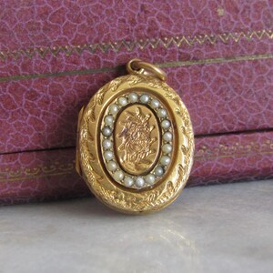 Op de afbeelding: Een goudkleurige ovale locket met een sierlijk ontwerp. De locket heeft een centrale ovaal met een bloemmotief, omringd door kleine parels. De buitenrand van de locket heeft een gedetailleerd, getextureerd patroon. De locket staat tegen een paarse achtergrond.
