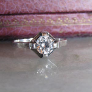 Peut inclure: Une bague en argent avec un gros diamant clair taillé en rond, serti dans un sertissage carré. Le corps de la bague est fin et simple, le diamant étant le point central. Il s'agit probablement d'une bague de fiançailles ou de caractère.
