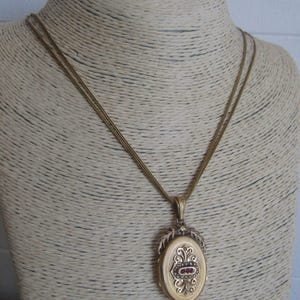 May include: A gold locket necklace with a detailed oval pendant featuring a red stone and intricate floral designs.