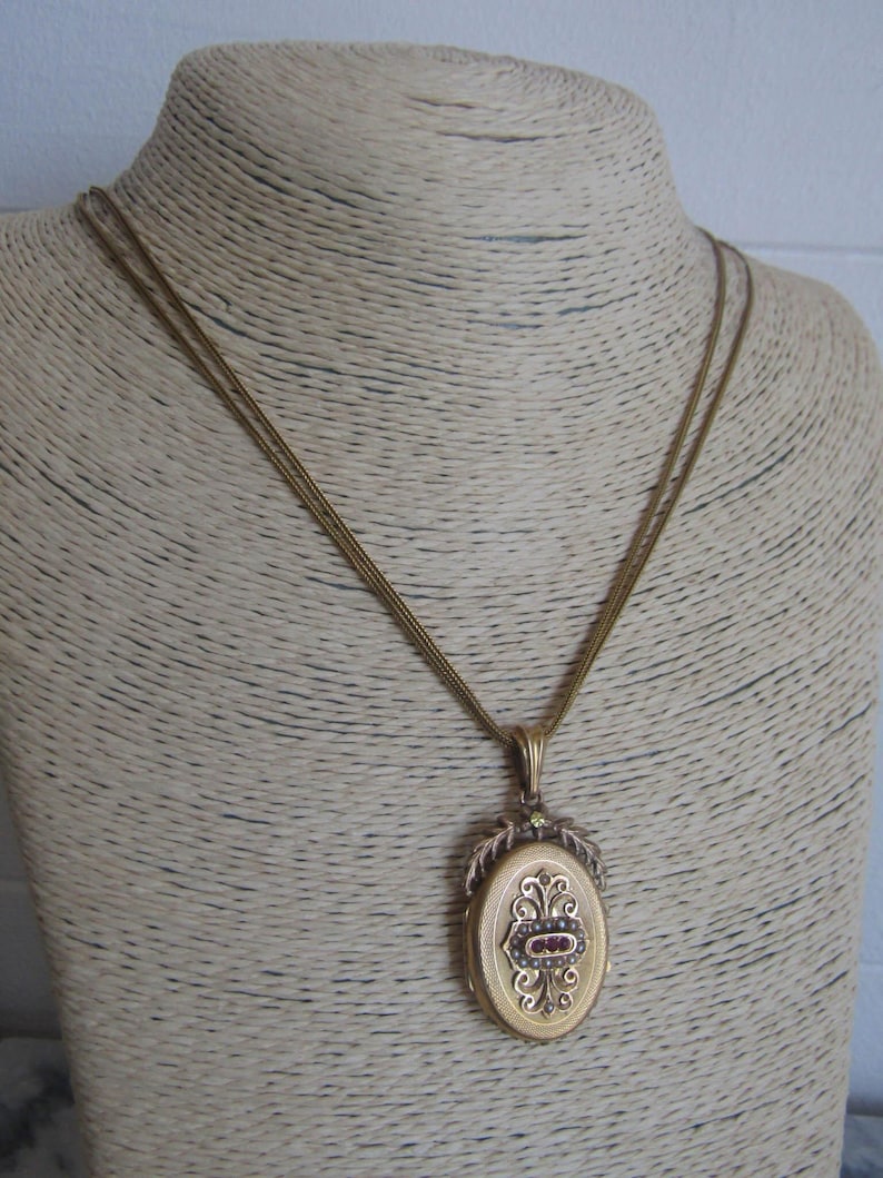 May include: A gold locket necklace with a detailed oval pendant featuring a red stone and intricate floral designs.