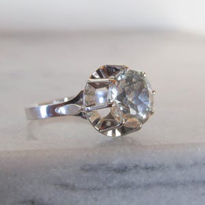 May include: A silver ring with a large, clear, round gemstone. The gemstone is set in a decorative, openwork setting. The ring band is smooth and polished, reflecting the light. The ring is displayed on a light-colored surface.