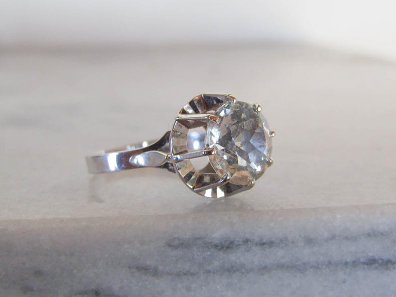 May include: A silver ring with a large, clear, round gemstone. The gemstone is set in a decorative, openwork setting. The ring band is smooth and polished, reflecting the light. The ring is displayed on a light-colored surface.